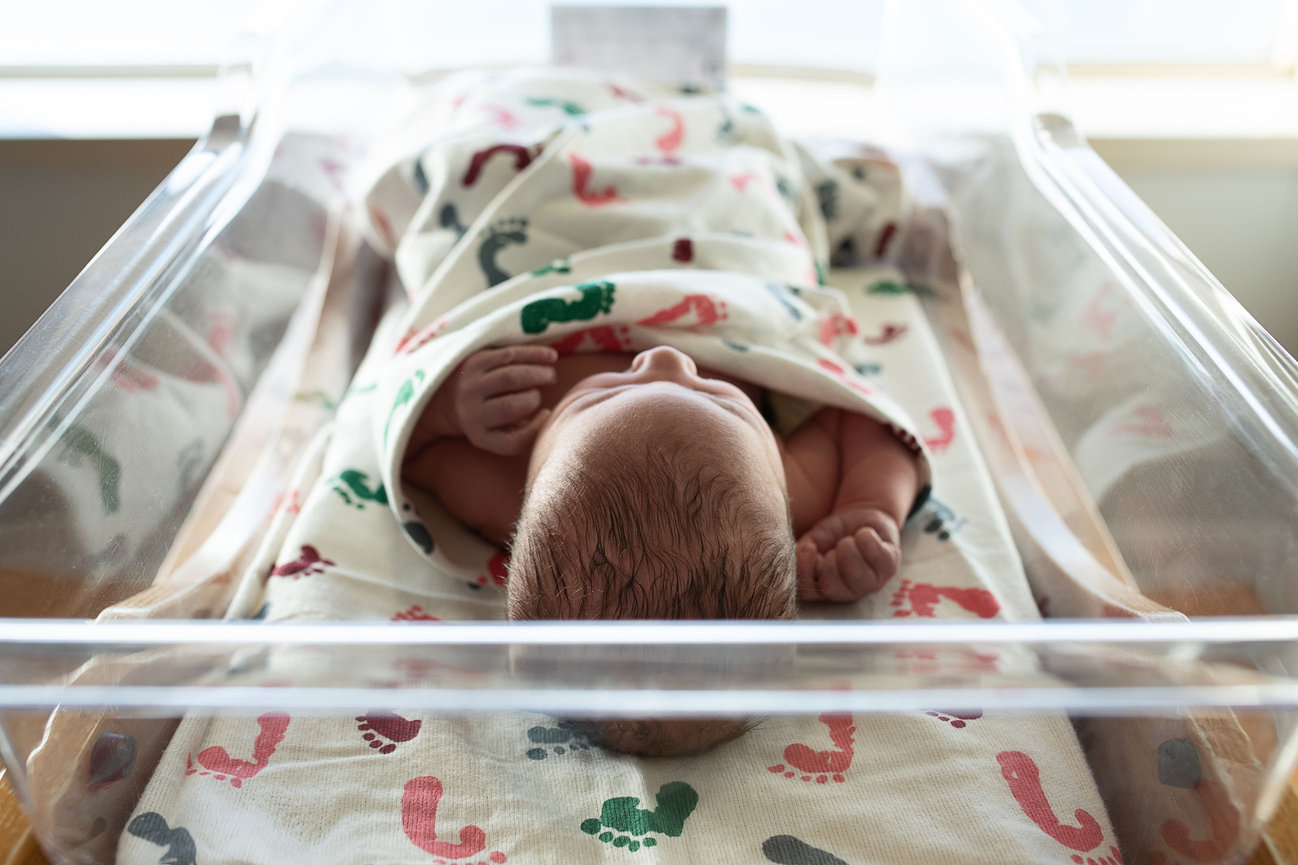 A newborn baby is swaddled in a hospital bassinet at Advocate Sherman Hospital in Elgin, wrapped in a white blanket with colorful footprints. The babys tiny hands are visible, and they lie under soft lighting.