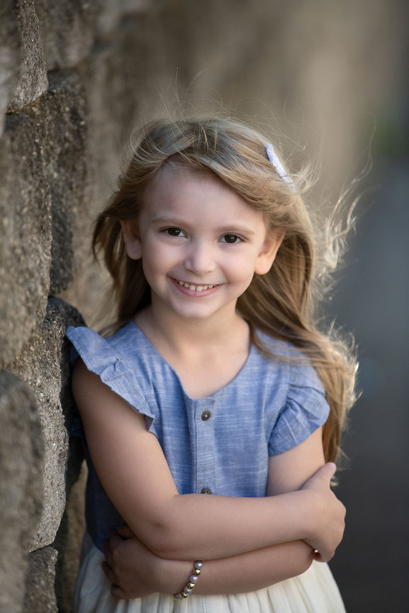 A young girl with long hair smiles at the camera, embodying the essence of child photography. She stands against a stone wall, wearing a blue top with ruffled sleeves and a cream skirt. The sunlight highlights her hair beautifully.