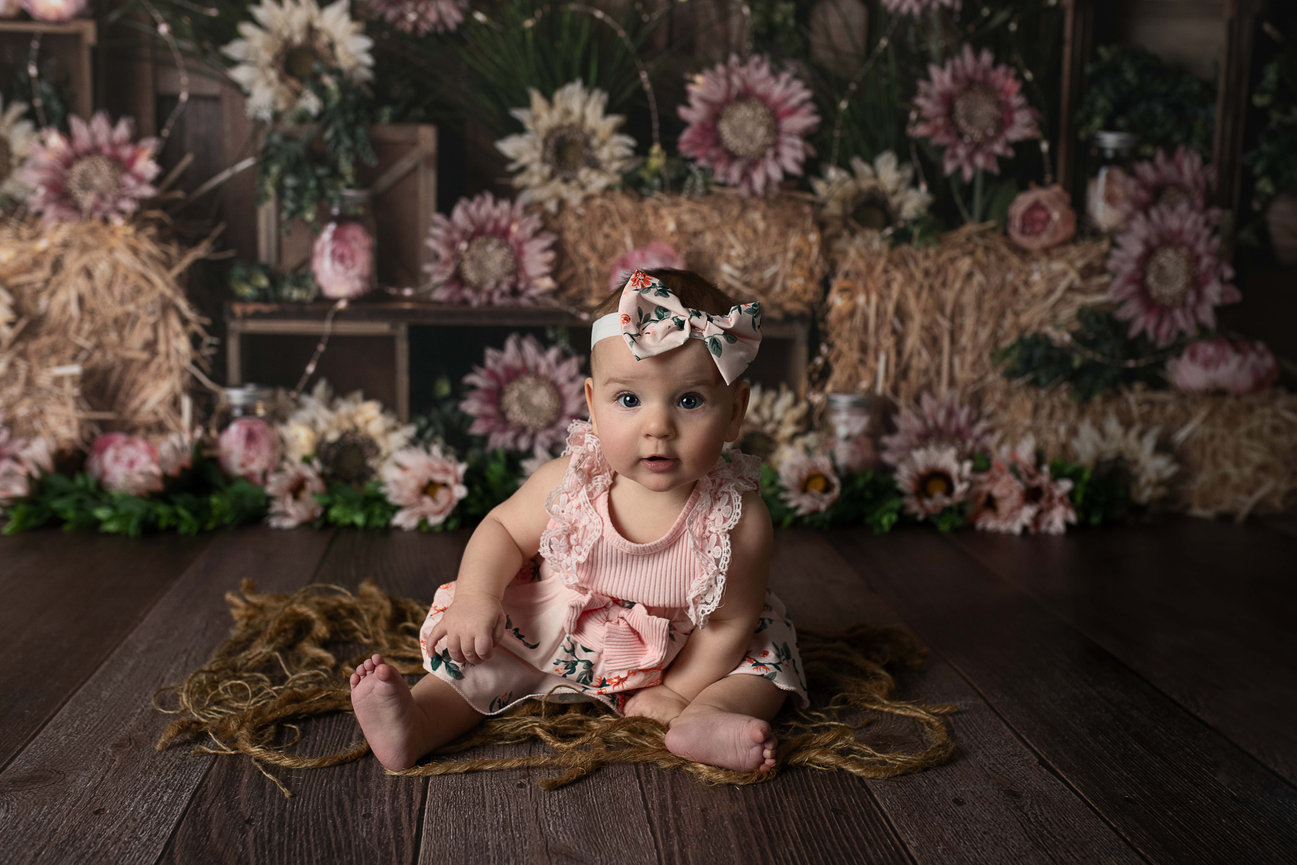 A newborn baby photo captures a baby wearing a floral headband and pink outfit as they sit on a rustic wooden floor. The background features an arrangement of pink and white flowers, straw, and greenery, creating a soft, whimsical setting.