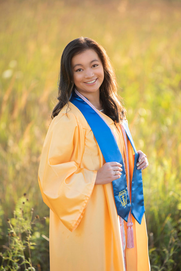 A person in a yellow graduation gown and blue stole smiles while standing in a sunlit field. They are looking towards the camera, and the background is softly focused with green and brown grass.