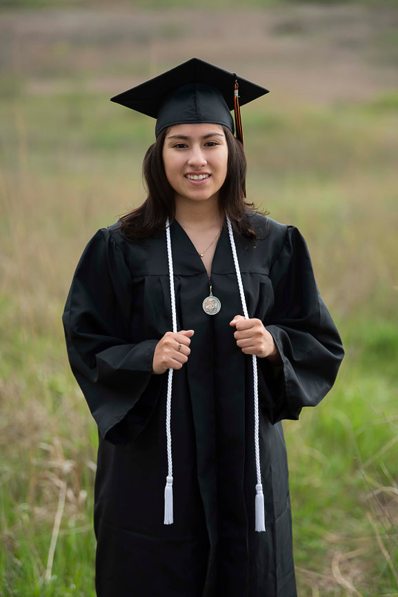 A person in a graduation cap and gown stands outdoors on a grassy field. They are smiling, holding their tassel draped over their gown, which is adorned with a white honor cord.
