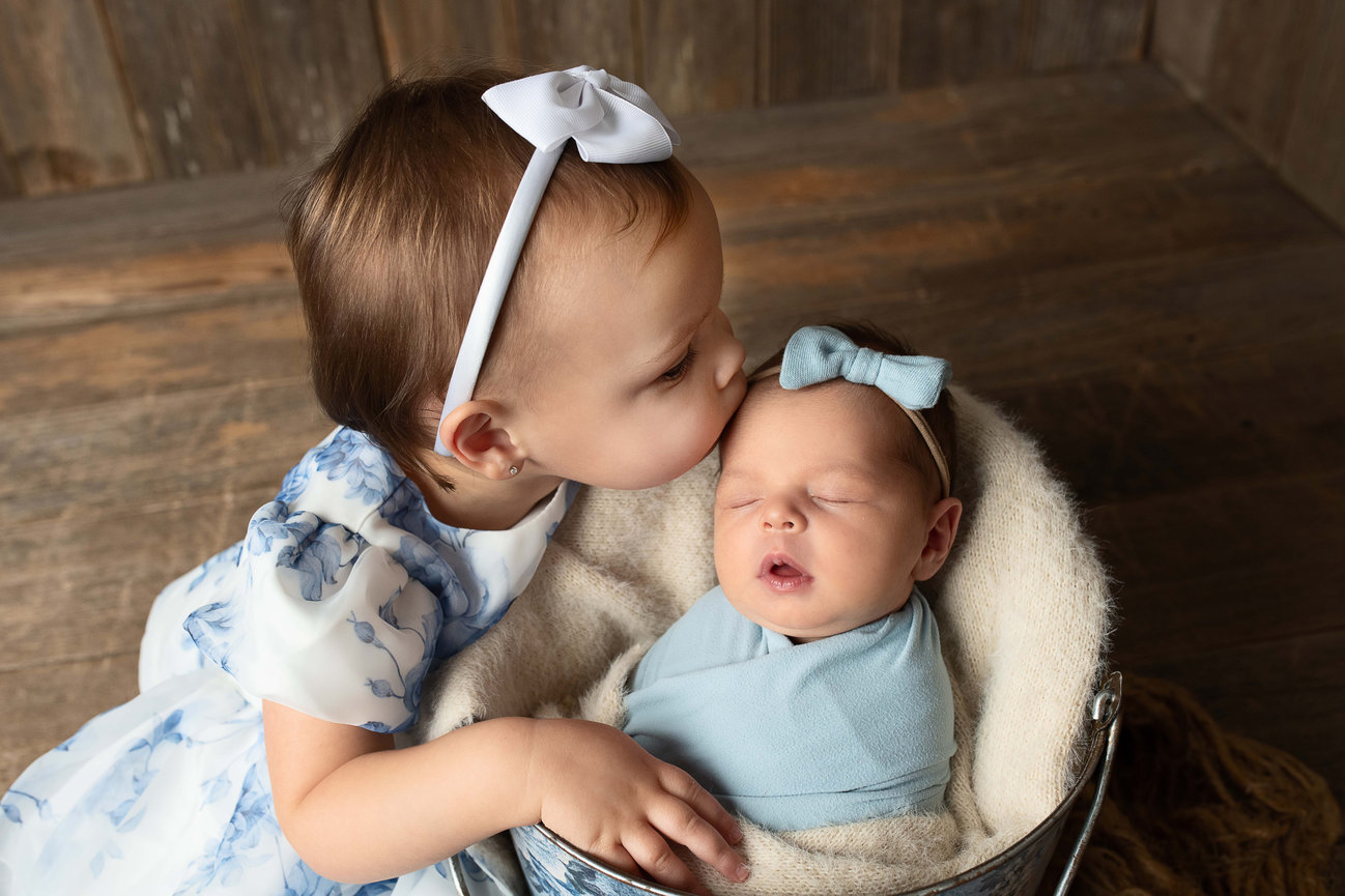 A toddler in a white and blue dress with a headband kisses a sleeping baby wrapped in a blue blanket and wearing a matching bow. The baby rests in a basket on a wooden floor.