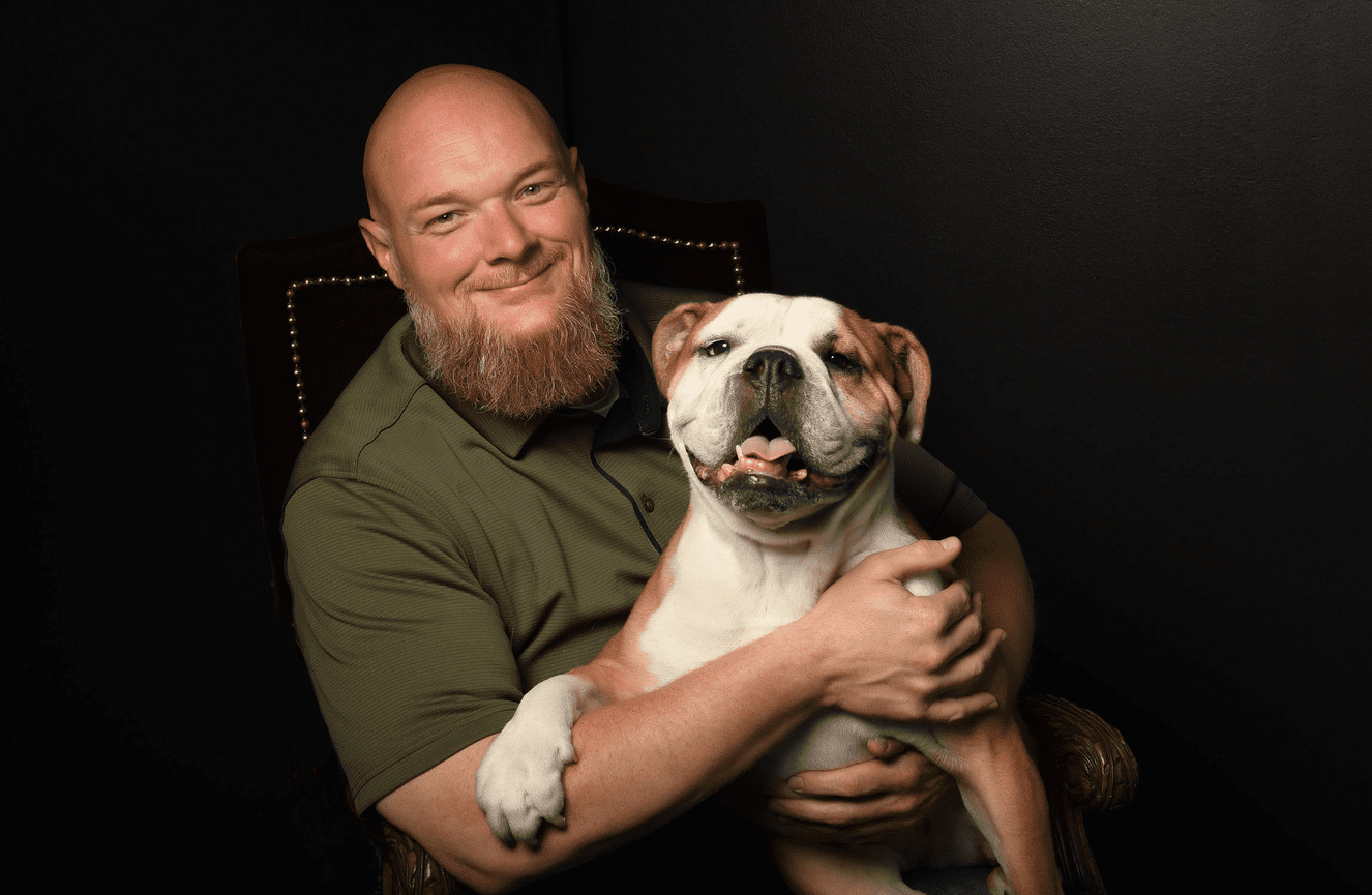 A bearded man in a green short sleeved shirt vegan named Billy Rebman sitting in a red velvet chair holding his English bulldog named Immy who had her paw on his arm in the Kliks Photography studio in Cedar Rapids Iowa.