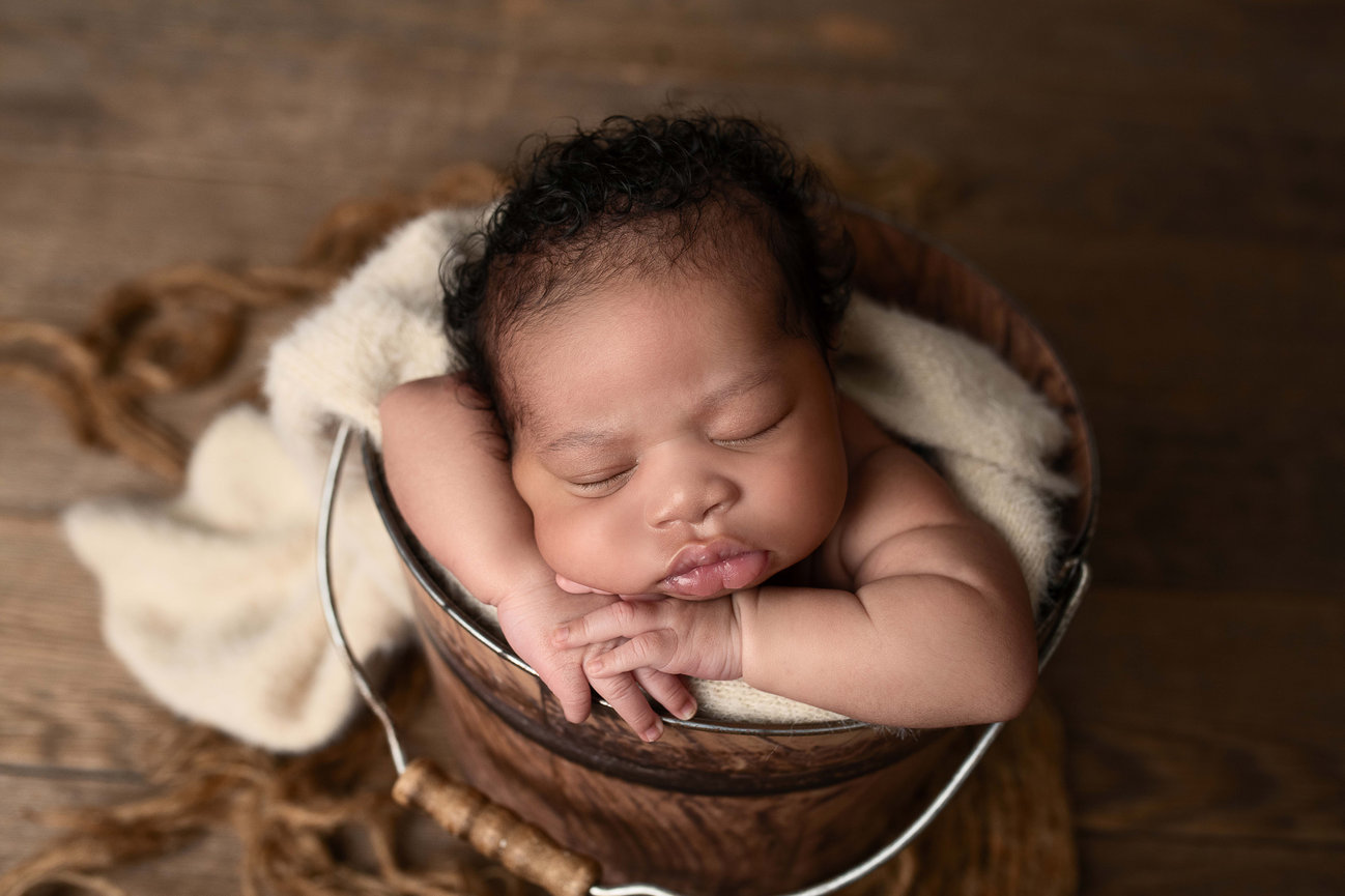 A newborn baby sleeps peacefully inside a wooden bucket, resting its head on folded arms. Wrapped in a soft cream blanket, the serene scene evokes the quiet charm of Almora. The background features a wooden floor and a coiled rope accent.