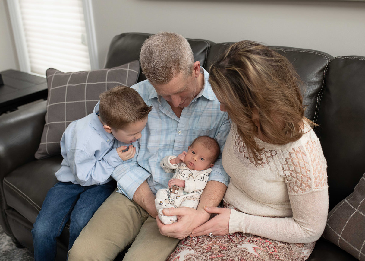 Family of four sitting on a couch, parents holding a newborn while a young boy leans in close