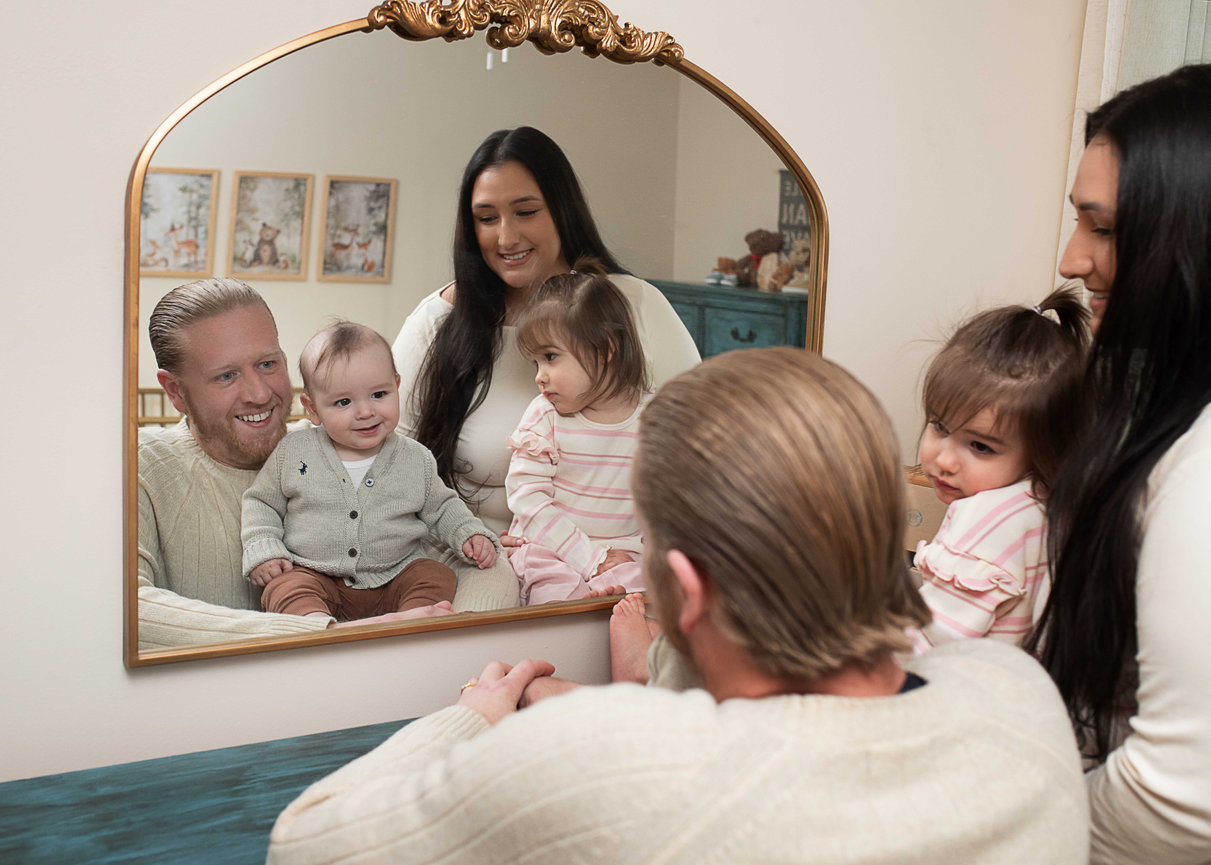 Family of four looking at themselves in a decorative mirror, with two toddlers and two adults smiling