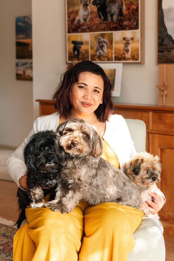 Ina Jalil sitting in an armchair in yellow trousers with three small dogs resting across her lap, framed dog portraits hanging on the wall behind her