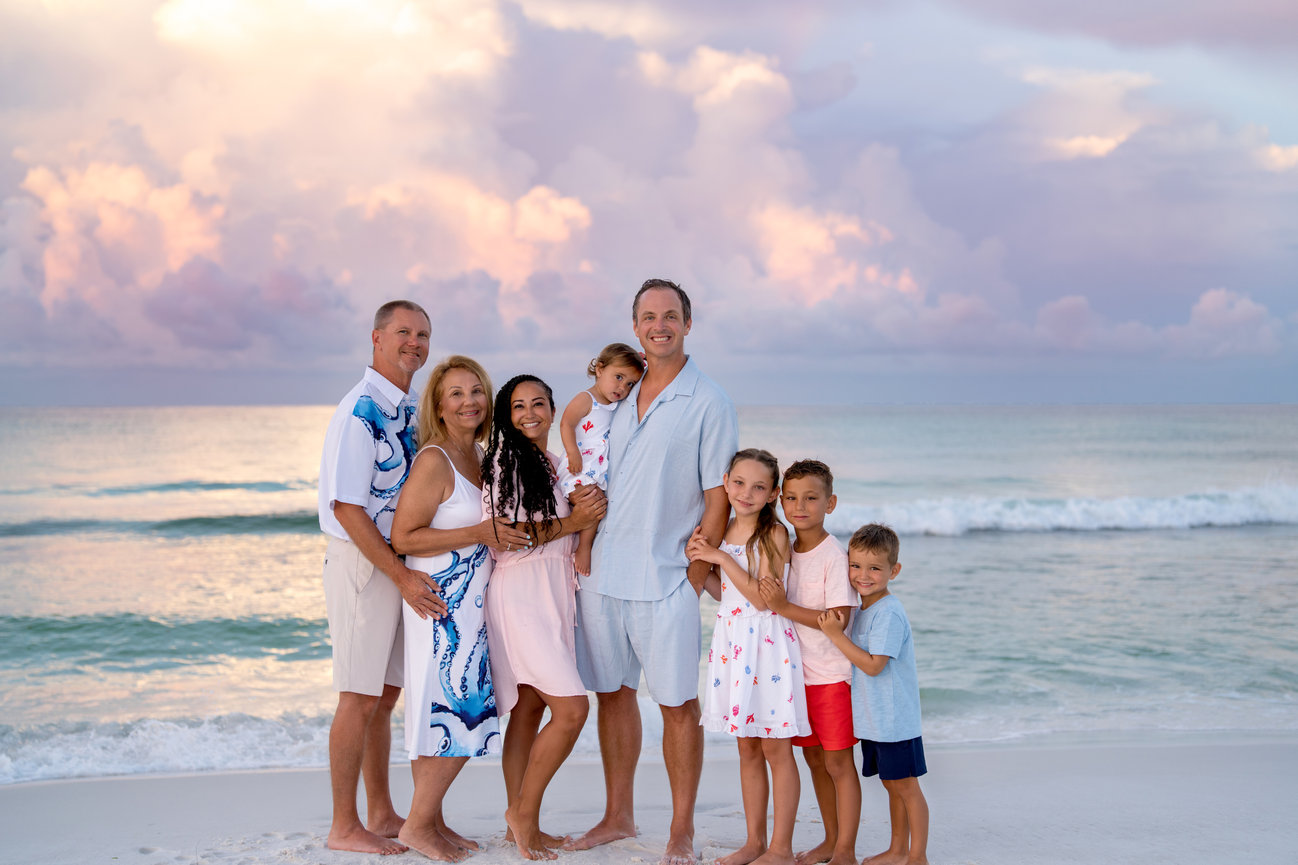 Family posing together on a beach at sunset, with pink clouds and gentle waves in the background.