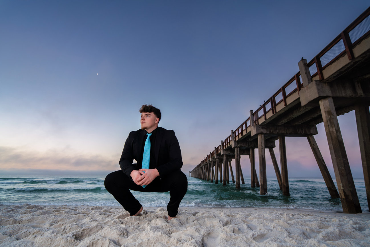 A person in a suit crouching on a sandy beach near an ocean pier at sunset.