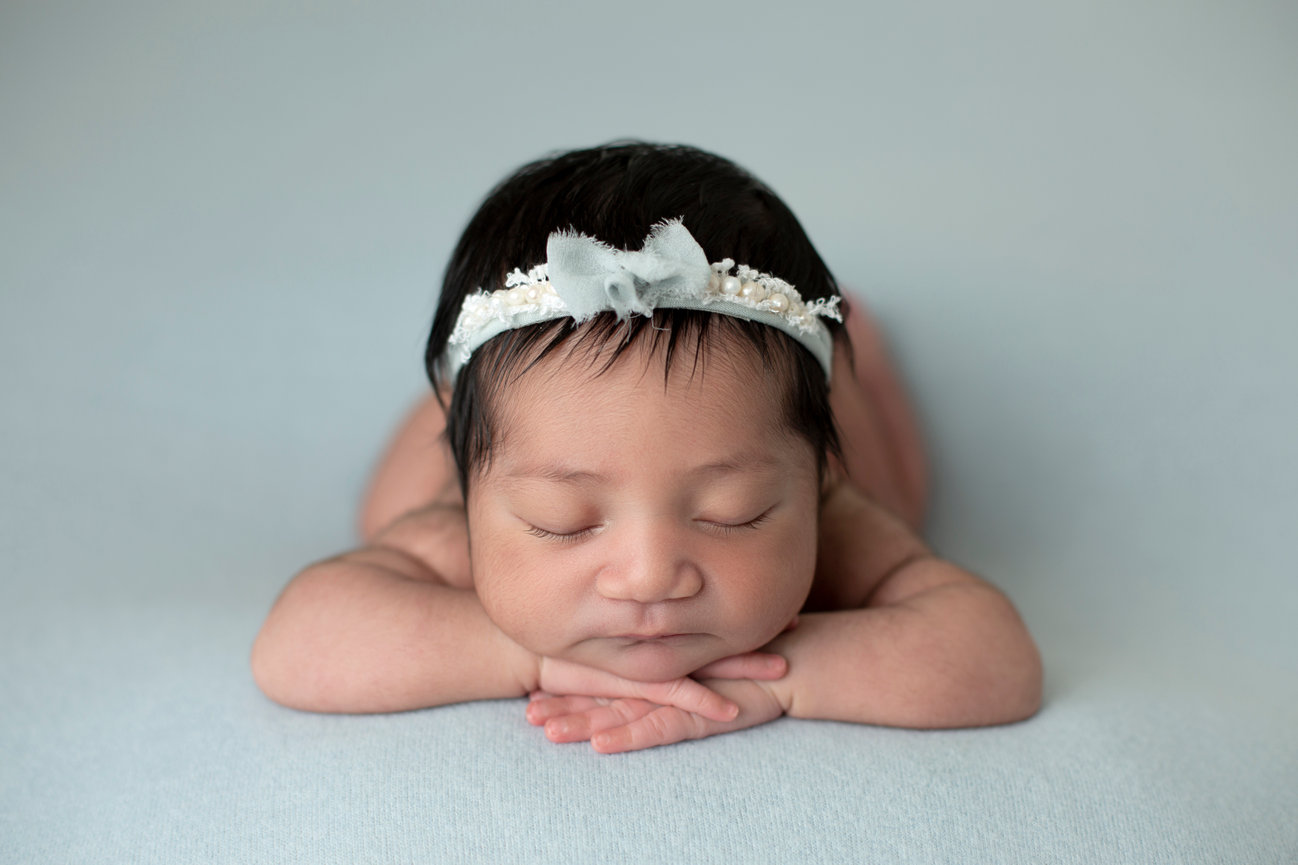 A posed newborn portrait of a baby girl with a thick head of hair. She is lying on a soft blue backdrop with her hands under her chin and wearing a headband with a blue bow, lace, and pearls.