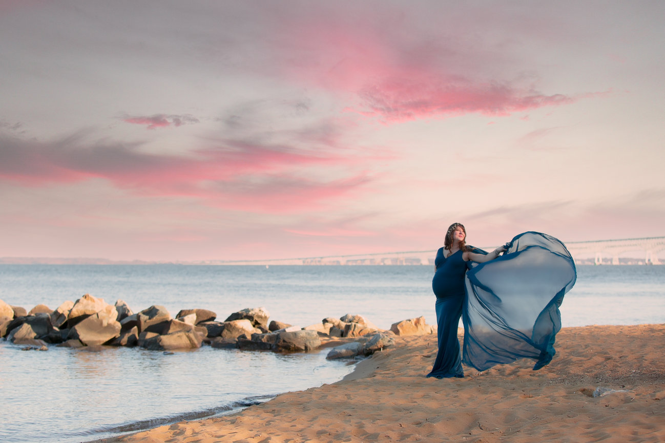 Dramatic Outdoor pregnancy portrait on a beach with rocks jutting out into the water, a bridge faintly seen in the background and fluffy pink clouds in the sky.  Momma is wearing a teal gown and the train of the gown is flowing in the wind.