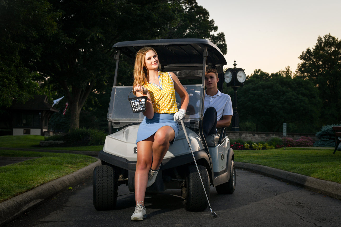 A woman in a yellow top and blue skirt poses confidently by a golf cart while a man sits inside, both on a golf course.