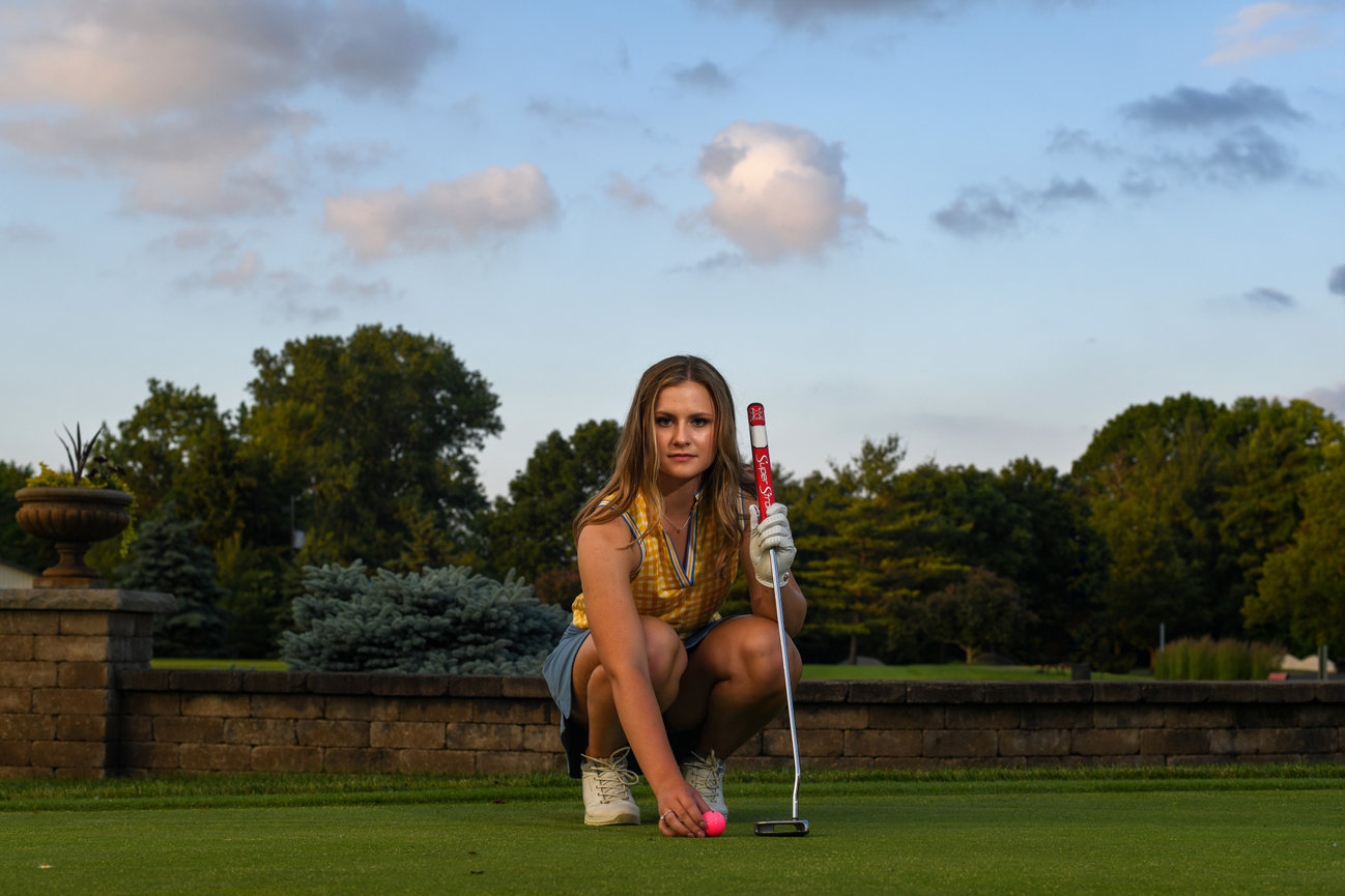Young woman preparing to putt on a golf course, wearing a yellow plaid top and denim shorts, with a pink golf ball.