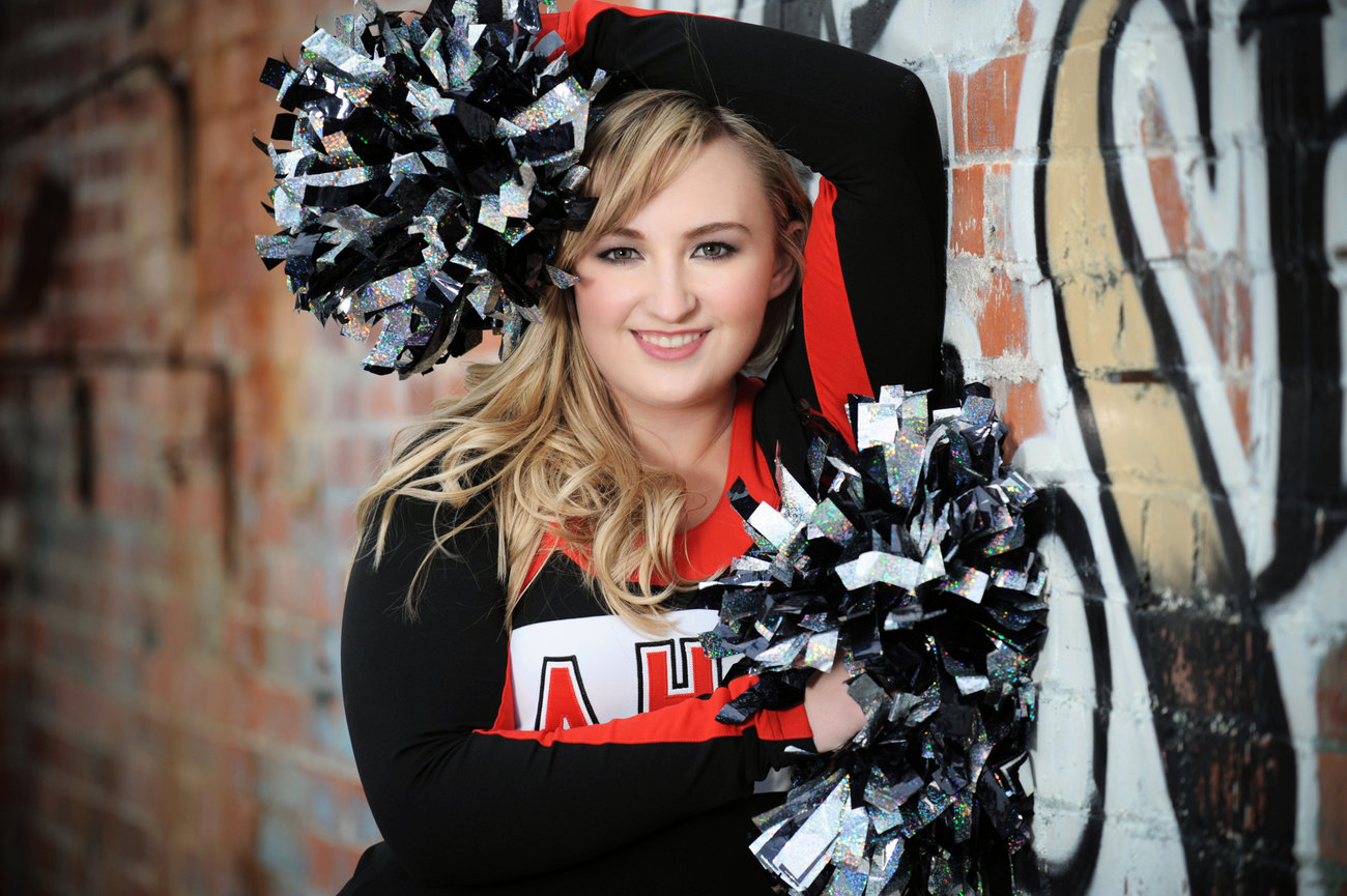 Cheerleader in a black and red uniform poses with pom-poms against a graffiti backdrop.