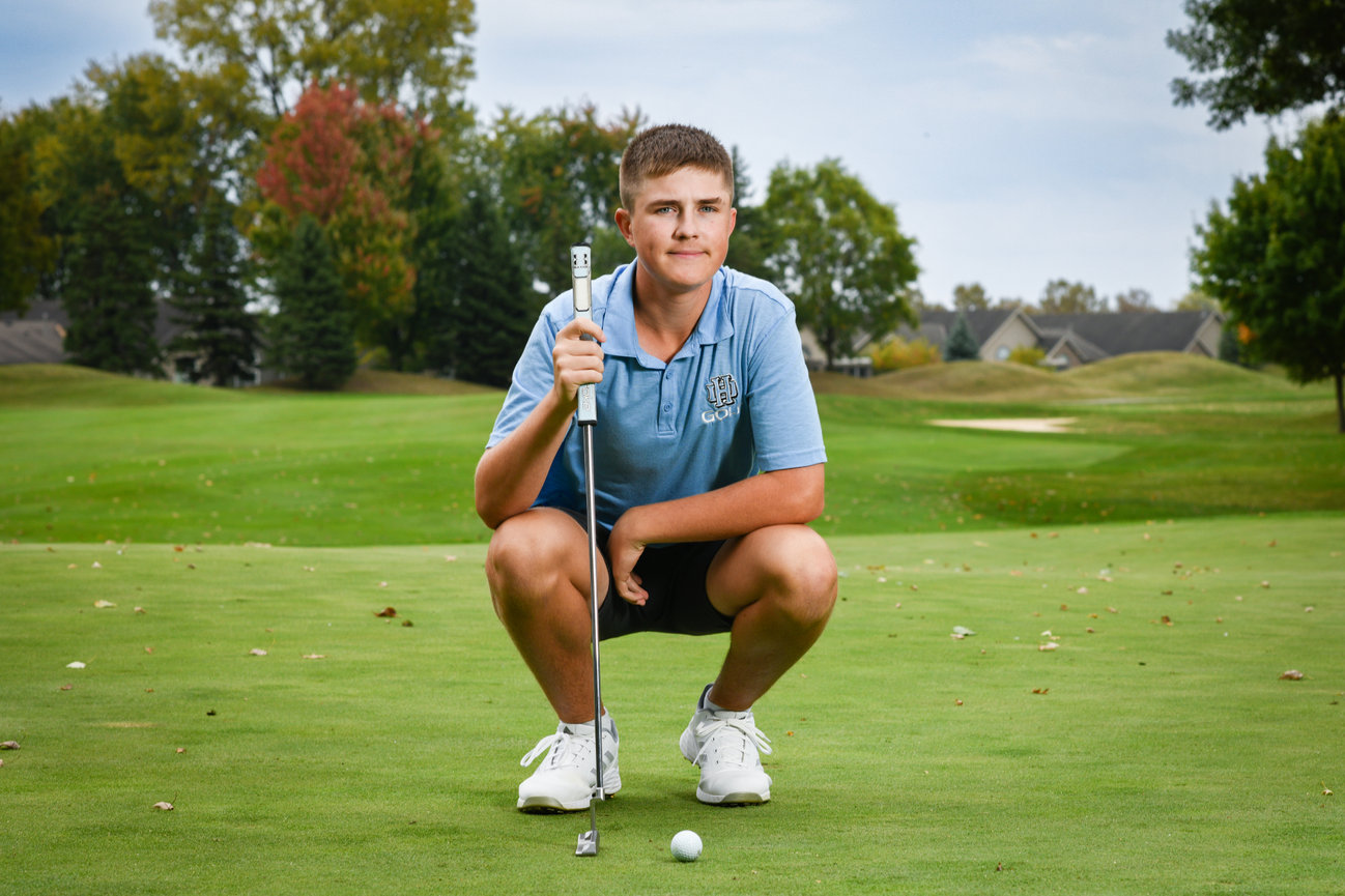 Young golfer crouching on a green, preparing to putt with a golf club in hand. Colorful trees in the background.