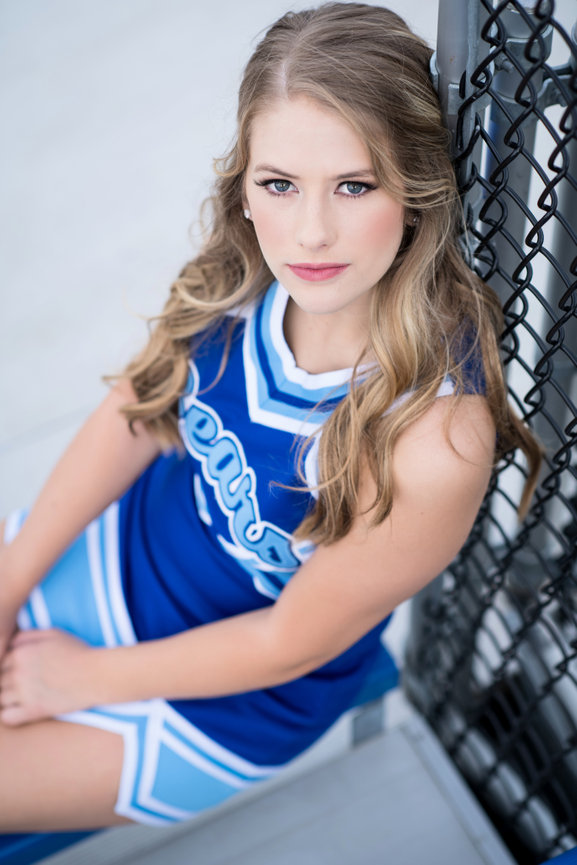 Cheerleader in a blue uniform sitting against a fence, looking confidently at the camera.