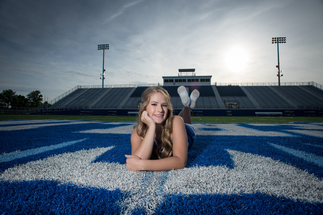 A young woman lies on a blue football field, smiling with stadium seats and a setting sun in the background.