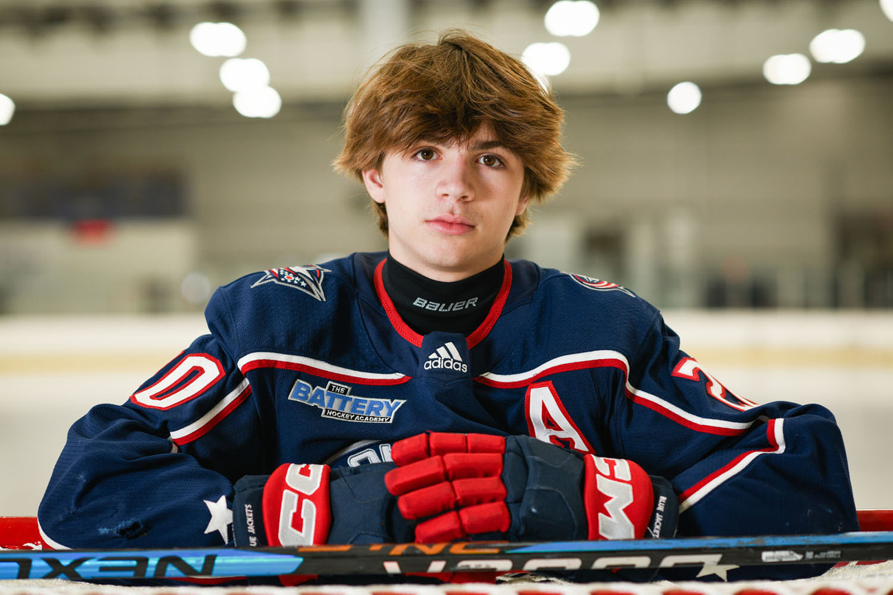 Young male hockey player in a blue jersey, resting his arms on a hockey stick, with an ice rink in the background.
