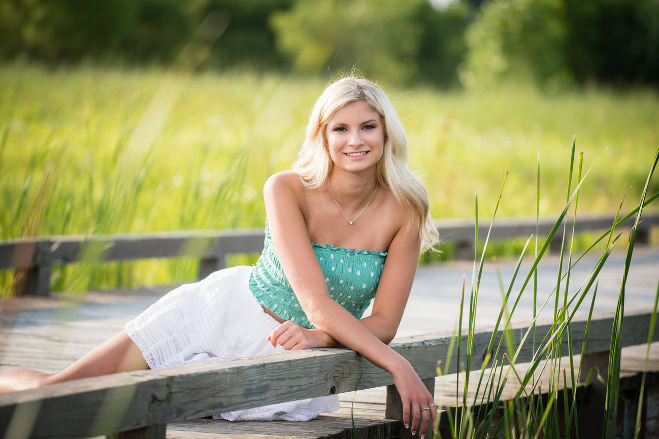 Blonde teenage girl in a green and white polka dot top and white skirt sitting on a bridge surrounded by grass for senior portraits in Dublin OH.