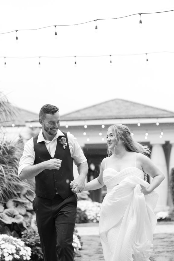 Bride and groom joyfully walk hand in hand under string lights in an outdoor setting.
